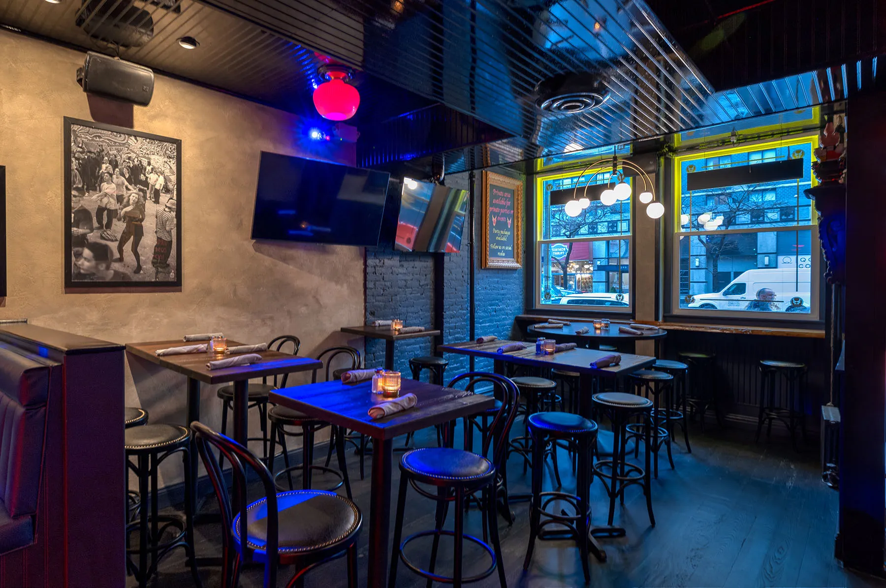 Sports bar lounge with HD TV, high-top tables and stools, framed vintage photo art, and street-facing windows with neon glow.
