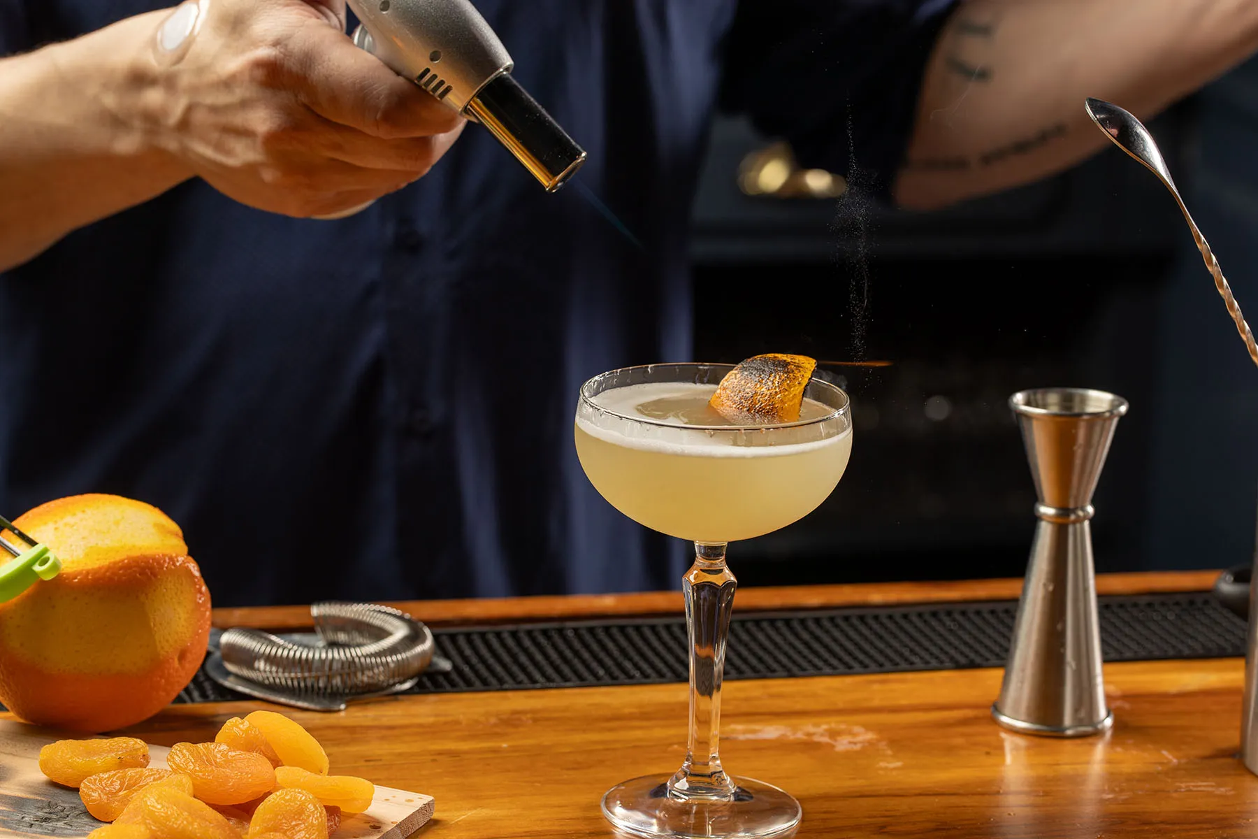 Bartender using a torch to add a smoky citrus garnish to a pale yellow craft cocktail.