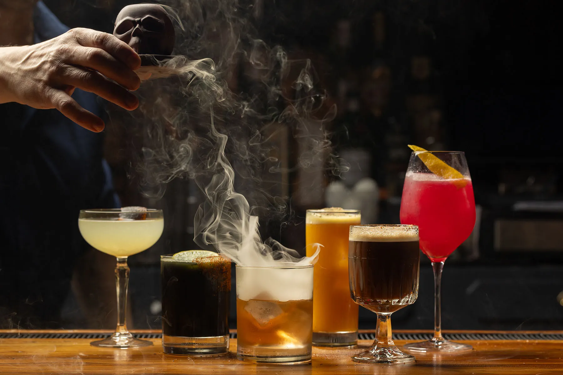 Assorted craft cocktails with a smoking whiskey drink and colorful garnishes.
