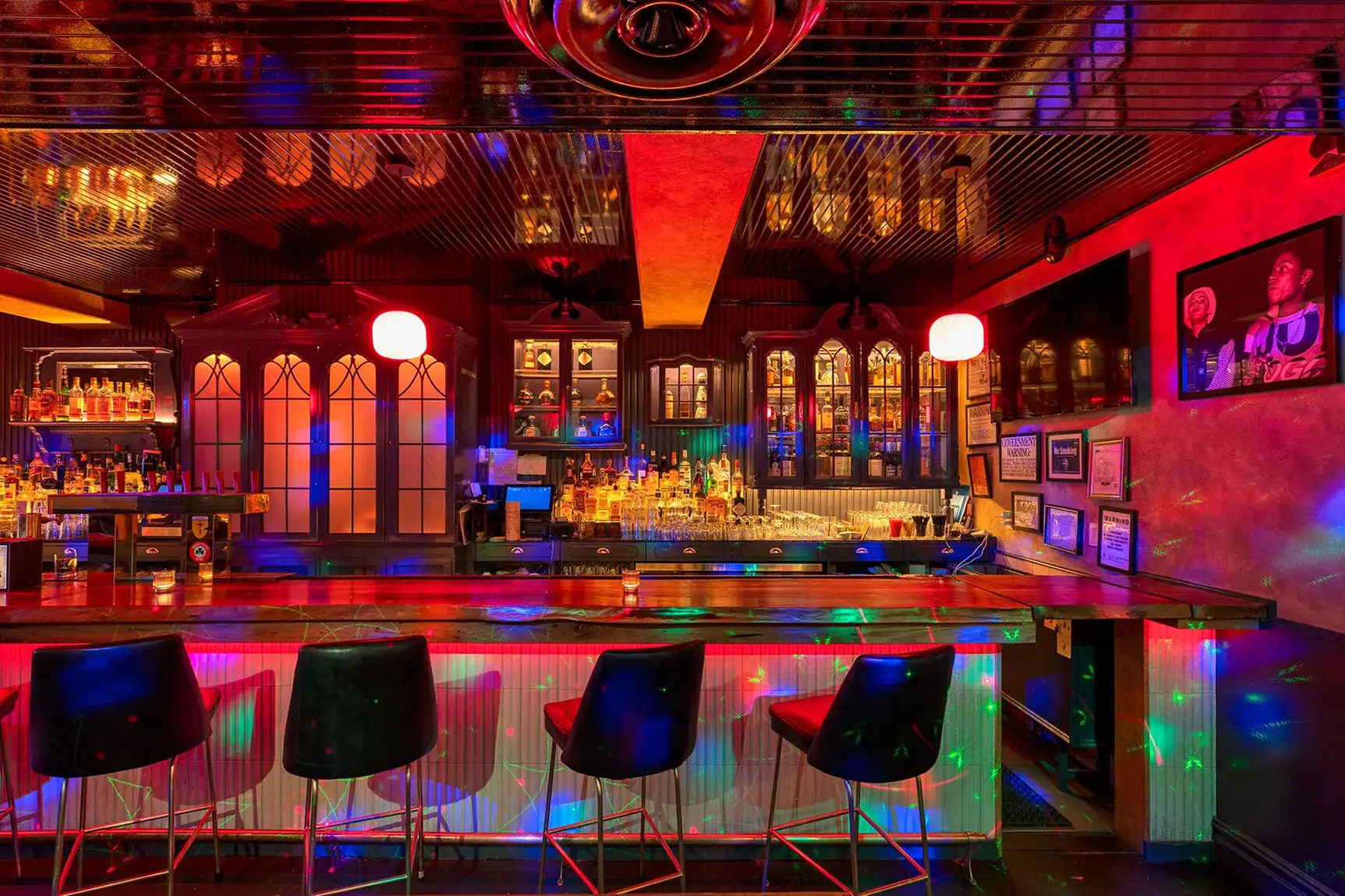 Vibrant bar interior with illuminated liquor shelves, black bar stools, red pendant lighting, and colorful disco-style light reflections on walls and floor.
