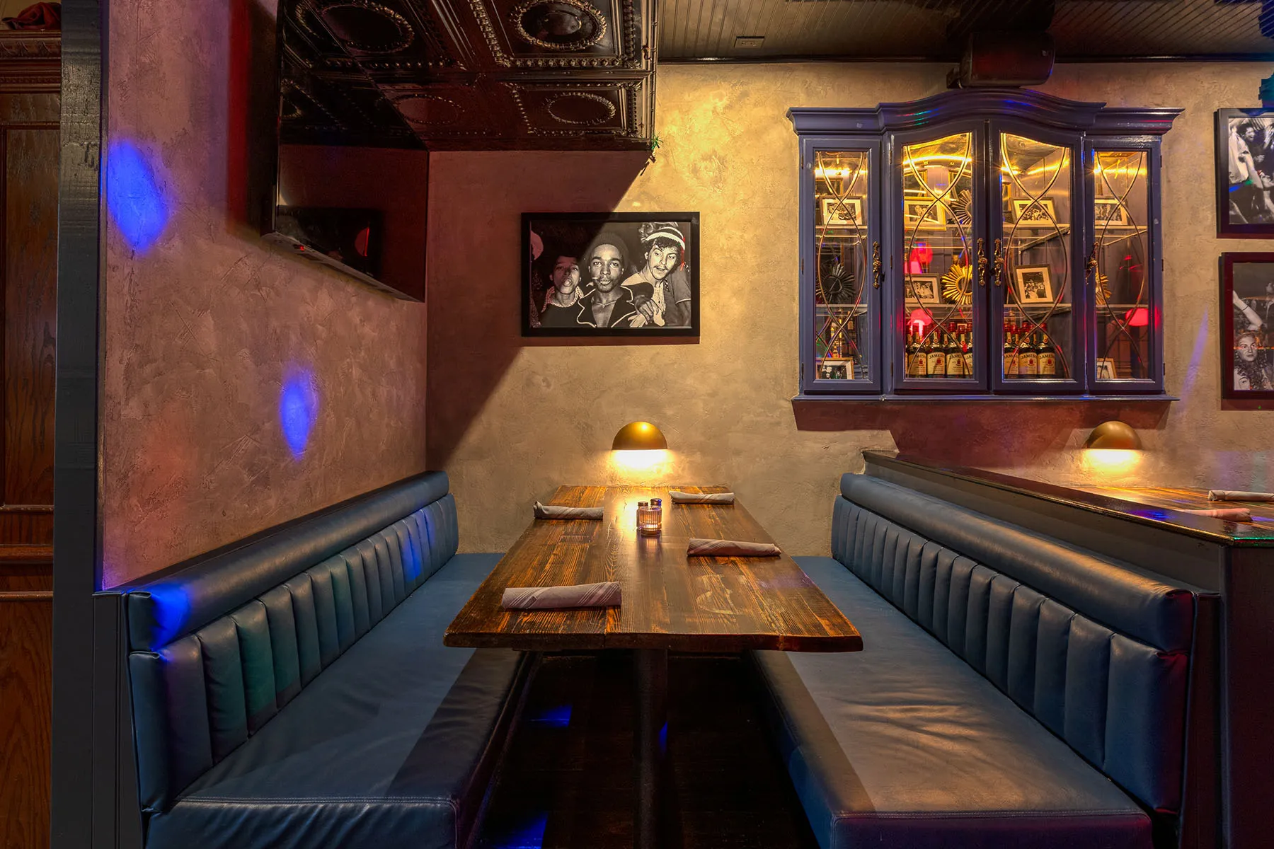 Cozy restaurant booth seating with dark wood table, blue leather benches, framed black-and-white photo, and backlit glass cabinet of bottles and photos.
