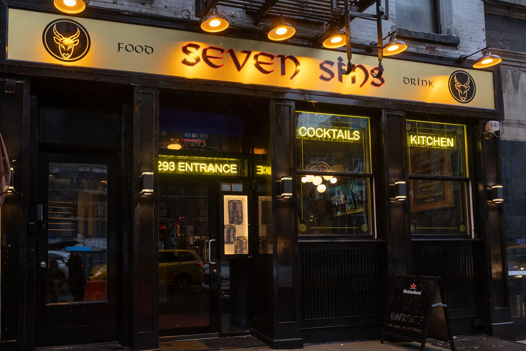 Street view of Seven Sins NYC sports bar with bright yellow signage and neon cocktail signs.