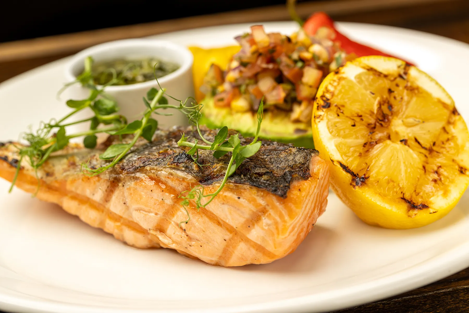 Grilled salmon fillet with charred lemon, avocado salsa, and herb garnish on a white plate.